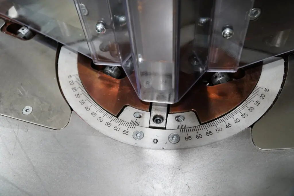Close-up of a circular degree scale on a metal machine base, showing measurements from 15 to 65, with a plastic safety guard positioned above the centre point