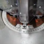 Close-up of a circular degree scale on a metal machine base, showing measurements from 15 to 65, with a plastic safety guard positioned above the centre point