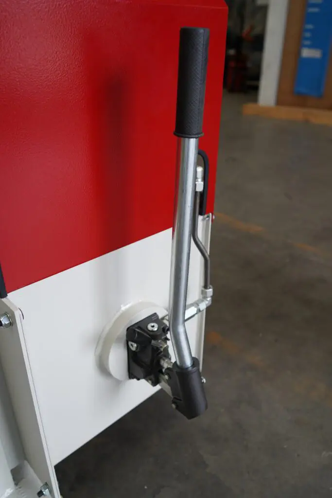Close-up of a manual hydraulic hand pump with a long silver lever and black grip, mounted to the side of a red and white industrial machine