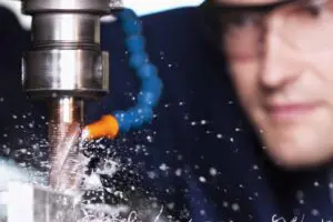 Close up image of a man wearing goggles watching a drill piece and coolant drilling into metal