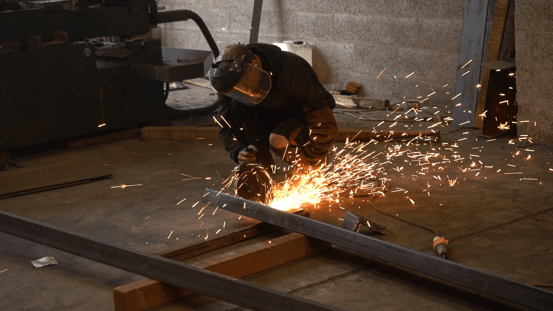 vlcsnap-2021-11-23-08h36m01s820 A worker wearing a safety mask uses an angle grinder to cut a metal beam on a workshop floor, creating a bright shower of sparks