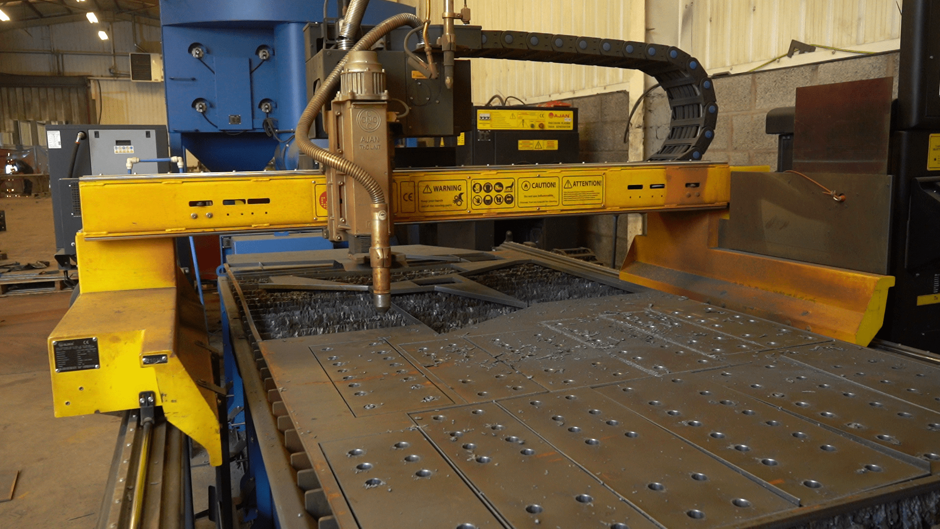 vlcsnap-2021-11-23-08h37m04s837 Ajan CNC plasma cutter with a yellow gantry and a torch head cutting precise shapes and holes into a large sheet of metal on a slatted cutting bed