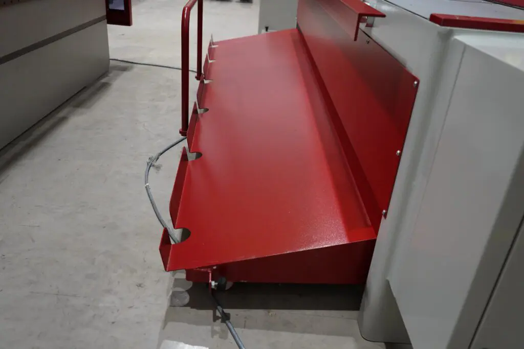 Close-up of an industrial machine base featuring two stacked, dark grey metal chip collection bins and a large tension spring